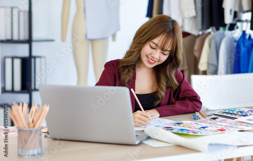 Millennial Asian young talent professional female dressmaker designer seamstress sitting working designing drawing sketching dress outline and choosing color pattern from collection at working desk.