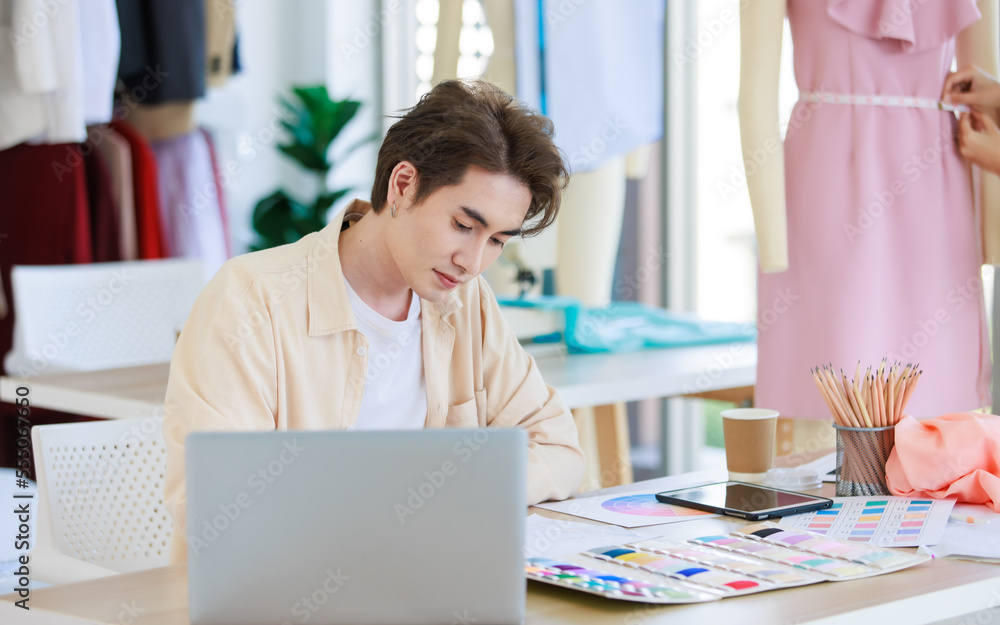 Millennial Asian young talent professional male dressmaker designer seamstress sitting working designing drawing sketching dress outline and choosing color pattern from collection at working desk Millennial Asian young talent professional male dressmaker designer seamstress sitting working designing drawing sketching dress outline and choosing color pattern from collection at working desk