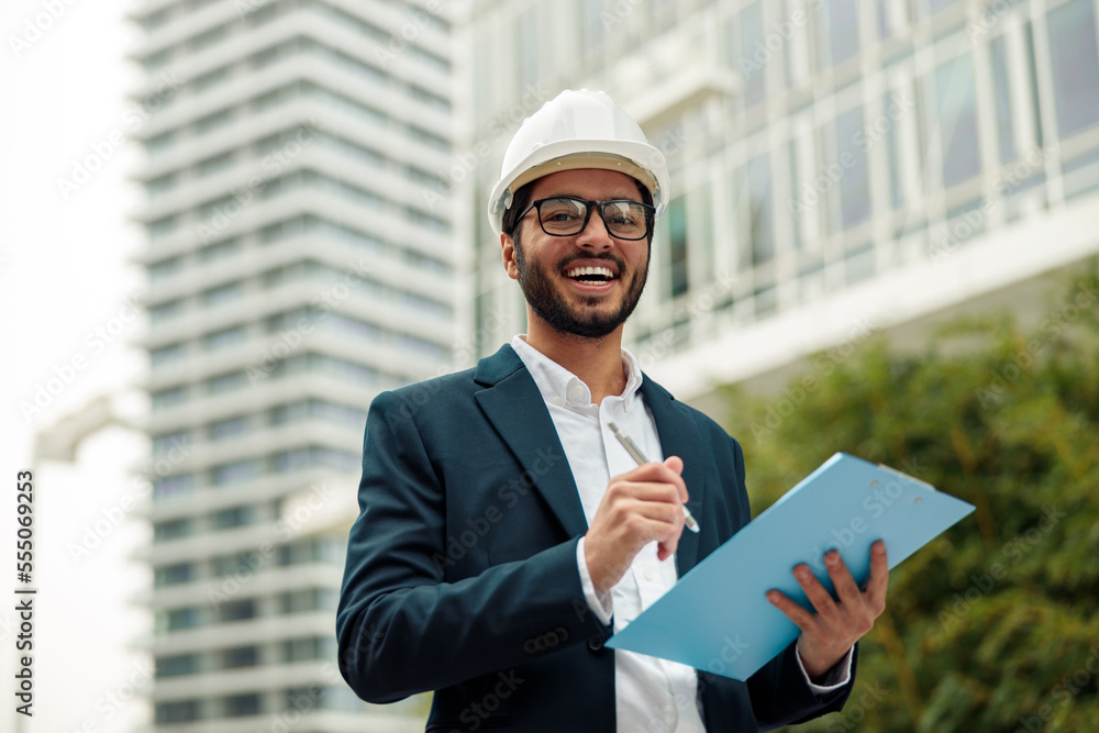 Indian construction architect in suit and hardhat making notes against modern building Indian construction architect in suit and hardhat making notes against modern building