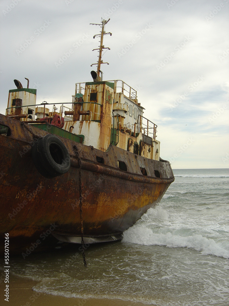 Fototapeta premium Old rusty tugboat on beach was run aground, stranded and abandoned, with bow in ocean waves in portrait view.