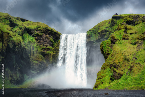 Majestic Skogafoss waterfall flowing with rainbow and moody sky in summer at Iceland