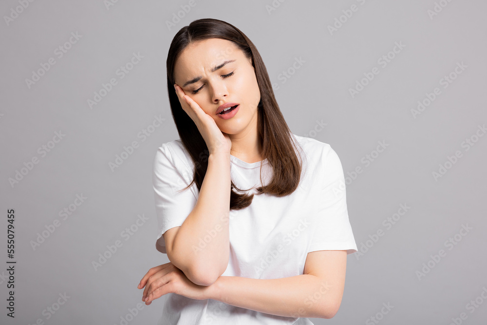 Sad exhausted young girl woman with headache suffering from migraine standing on grey background in studio isolated closing eyes holding arm near head. Sad exhausted young girl woman with headache suffering from migraine standing on grey background in studio isolated closing eyes holding arm near head.