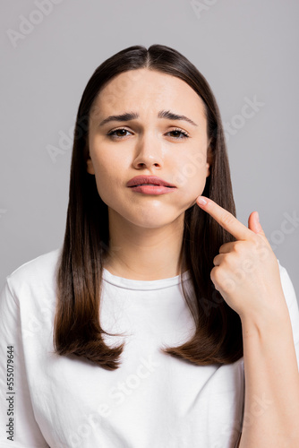 Crying tired sad woman suffers from painful toothache dental illness pointing on teeth standing on grey background in studio isolated wearing basic white t-shirt.