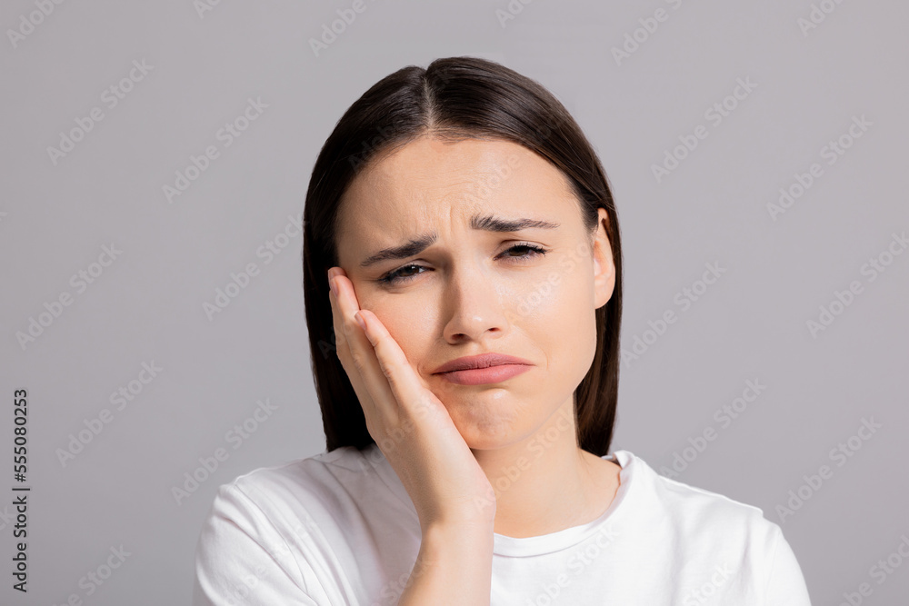 Sad exhausted tired young woman suffering from toothache pain standing on grey background in studio isolated wearing white basic t-shirt having dental illness waiting for dentist appointment. Sad exhausted tired young woman suffering from toothache pain standing on grey background in studio isolated wearing white basic t-shirt having dental illness waiting for dentist appointment.