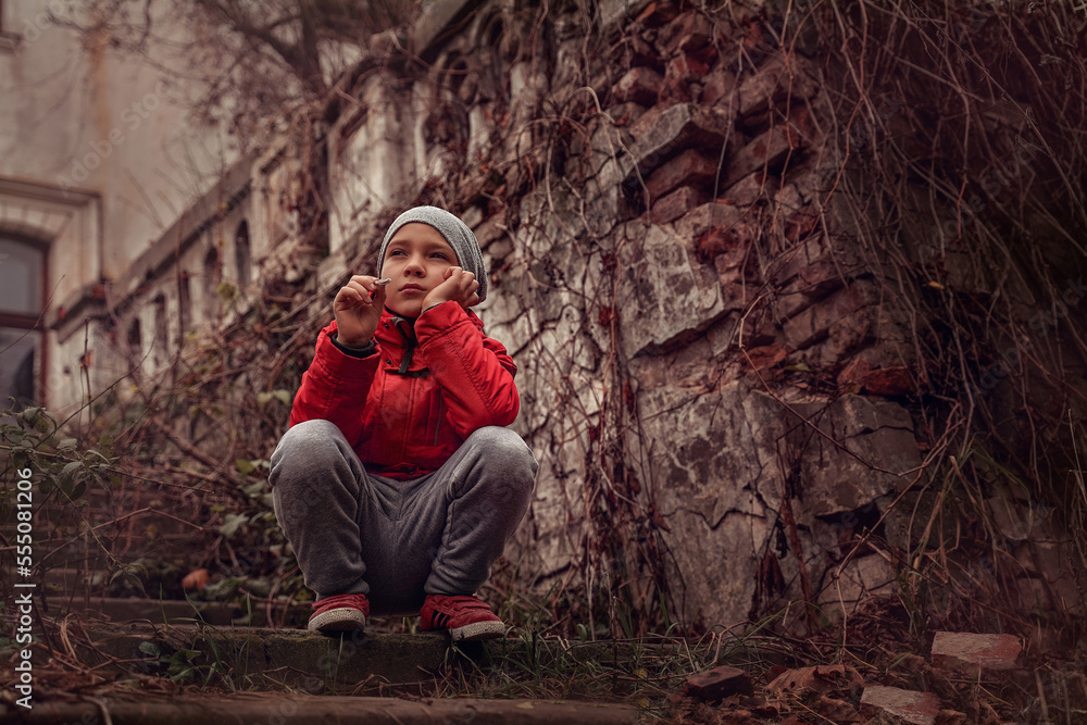 Sad guy in autumn sits on steps against background of building ruins, around dried shoots of plants, fallen foliage. Boy looks thoughtfully into the sky, his head propped up with hand, view from below