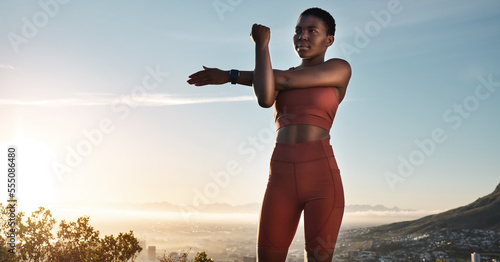 Fitness, black woman and stretching arm on mountains, blue sky or sunset for wellness, healthy goals or energy. Focus, athlete and sports warm up, exercise and start body training with strong mindset photo