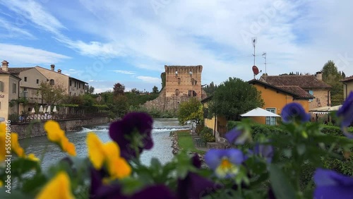 Scenic view through yellow and blue colorful flowers of Borghetto sul Mincio charming ancient Italian hamlet in Veneto, Italy