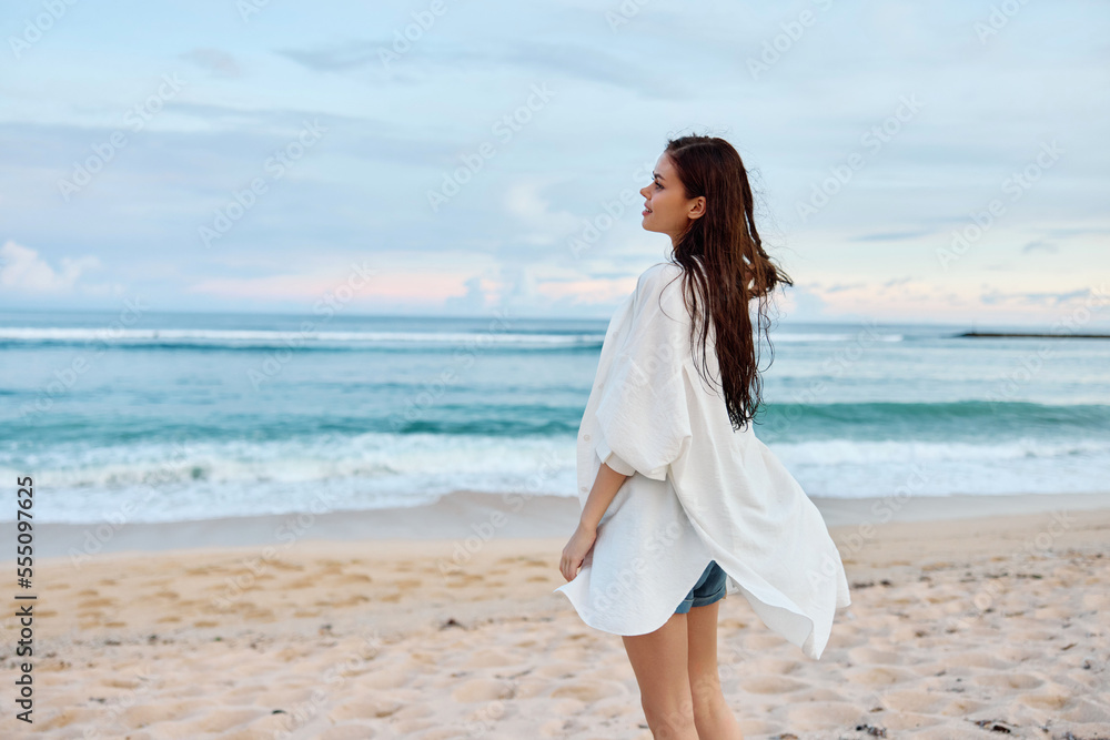 Happy tanned slim woman in white swimsuit shirt and jean shorts walks on the beach on the sand by the ocean with wet hair after swimming, sunset light Happy tanned slim woman in white swimsuit shirt and jean shorts walks on the beach on the sand by the ocean with wet hair after swimming, sunset light