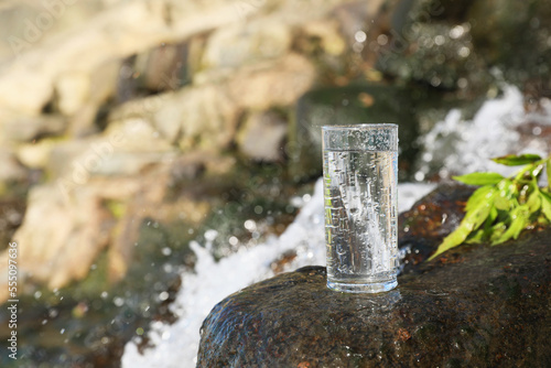 Wet glass of water on rocks near flowing stream outdoors, space for text