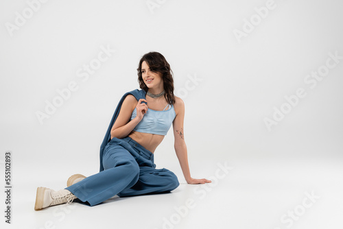 tattooed woman in blue trousers and crop top sitting with jacket and looking away on grey background