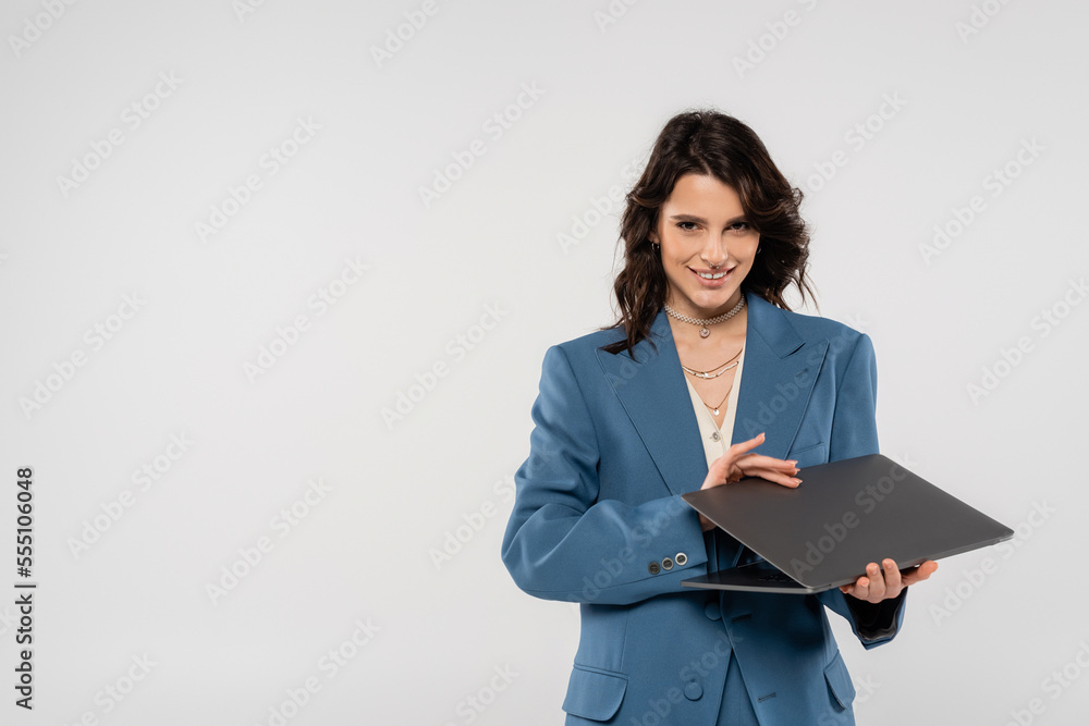 happy brunette woman in blue blazer holding laptop and looking at camera isolated on grey happy brunette woman in blue blazer holding laptop and looking at camera isolated on grey