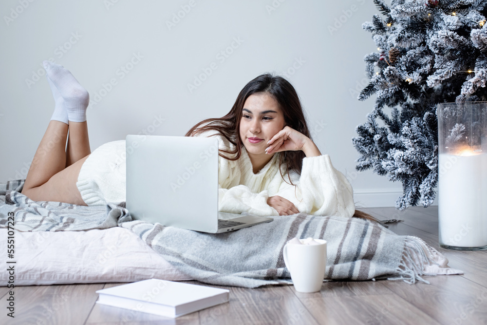 Woman in warm white winter sweater lying in bed at home using laptop Woman in warm white winter sweater lying in bed at home using laptop