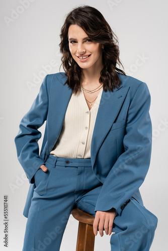 stylish brunette woman holding hand in pocket of blue pants and smiling at camera isolated on grey