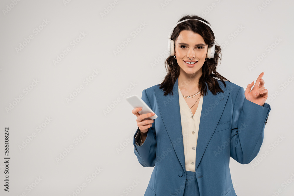 pleased brunette woman in wireless headphones holding smartphone and pointing with finger isolated on grey pleased brunette woman in wireless headphones holding smartphone and pointing with finger isolated on grey