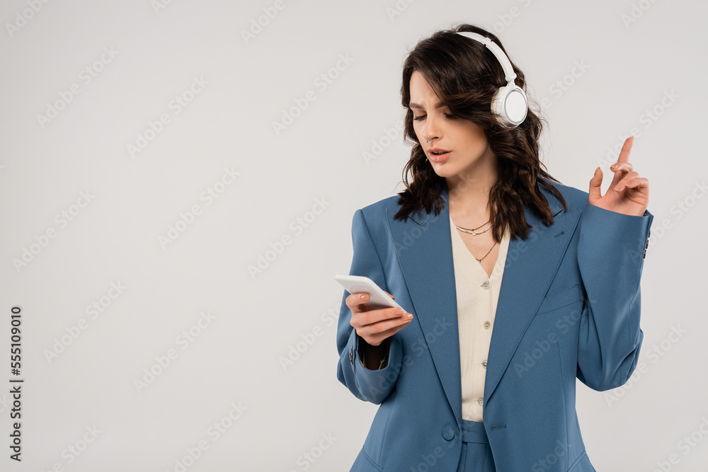 trendy brunette woman in wireless headphones looking at smartphone and pointing with finger isolated on grey trendy brunette woman in wireless headphones looking at smartphone and pointing with finger isolated on grey