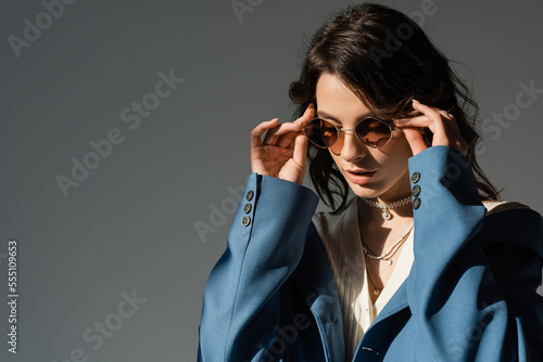 trendy woman in blue blazer and necklaces adjusting sunglasses isolated on grey