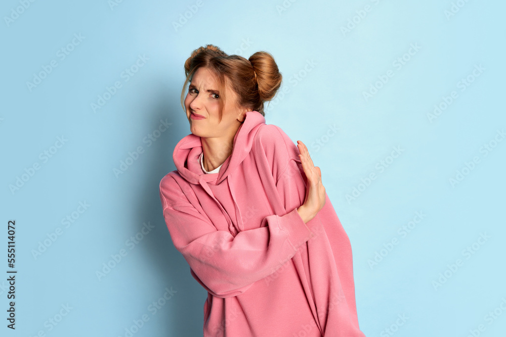 Portrait of young girl in pink hoodie posing with unpleasant face over blue studio background. Refusal, rejection gesture Portrait of young girl in pink hoodie posing with unpleasant face over blue studio background. Refusal, rejection gesture