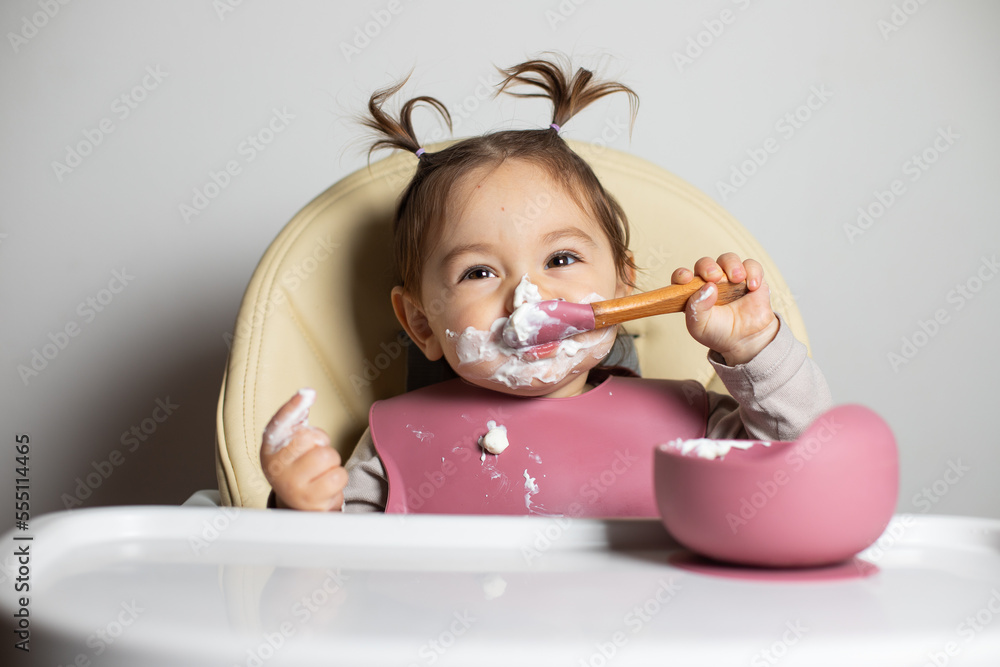 Small cute little toddler brunette caucasian girl with two tails tasting and enjoying by herself with a spoon the greek yogurt sitting in baby chair with messy face; self-feeding concept Small cute little toddler brunette caucasian girl with two tails tasting and enjoying by herself with a spoon the greek yogurt sitting in baby chair with messy face; self-feeding concept