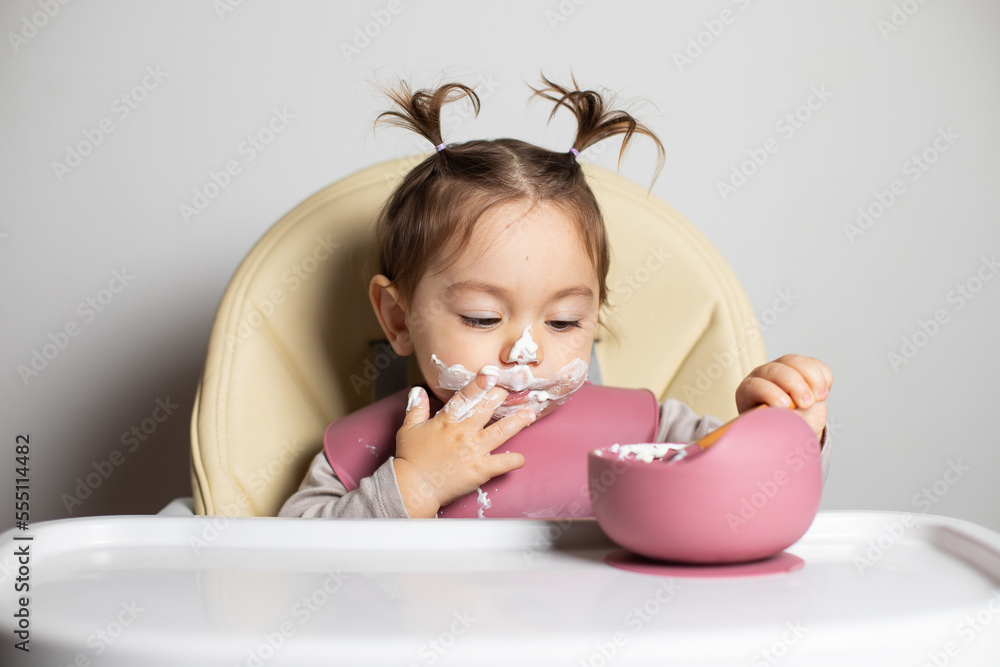 Small cute little toddler brunette caucasian girl with two tails tasting by herself with a spoon the greek yogurt sitting in baby chair with messy face; self-feeding concept Small cute little toddler brunette caucasian girl with two tails tasting by herself with a spoon the greek yogurt sitting in baby chair with messy face; self-feeding concept