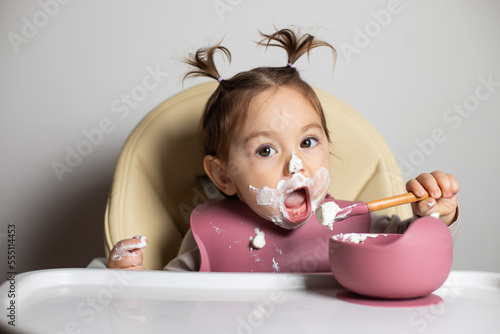 Small cute little toddler brunette caucasian girl with two tails eating by herself with a spoon the greek yogurt sitting in baby chair; self-feeding concept photo