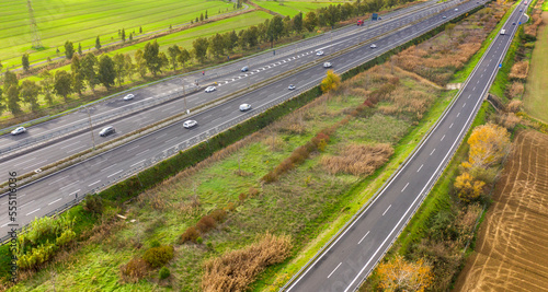 Aerial view of a stretch of highway in the Italian countryside. The road is divided into many lanes in each direction. There are cars and trucks on the street.Around the road there are trees and grass