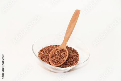 Small grains of natural brown buckwheat in a transparent glass plate and in a wooden spoon on a white background