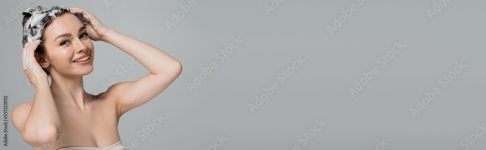 Naklejka premium cheerful young woman washing wet and foamy hair isolated on grey, banner