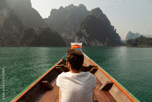 A young man, a backpacker, looks out from the bow of a longtail boat over the landscape and rainforest at Cheow Lan Lake, Khao Sok National Park
