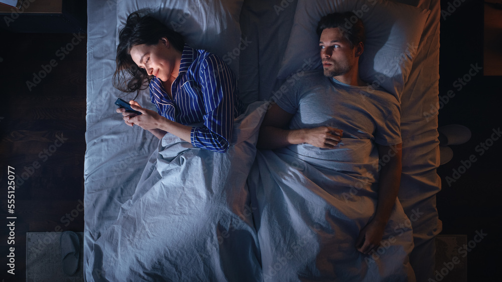 Top View Bedroom Apartment: Young Woman Uses Smartphone in Bed at Night When Her Male Partner Trying to Fall Asleep Beside. Couple Fight, Argue. Social Media, Doom Scrolling, Fake News Addiction Top View Bedroom Apartment: Young Woman Uses Smartphone in Bed at Night When Her Male Partner Trying to Fall Asleep Beside. Couple Fight, Argue. Social Media, Doom Scrolling, Fake News Addiction