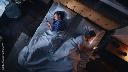 Top View Apartment Bedroom: Caucasian Young Couple Lying in Bed, Using Smartphones. Family of Two Using Mobile Phones to Browse Social Media, Search internet, Online Shopping Before Sleep. © Gorodenkoff