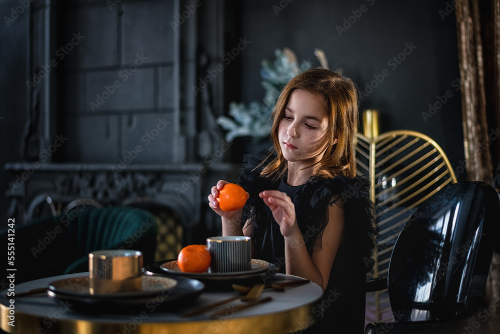 a cute elegant girl in a black dress sits at a table and holds a tangerine in her hand. holiday, new year