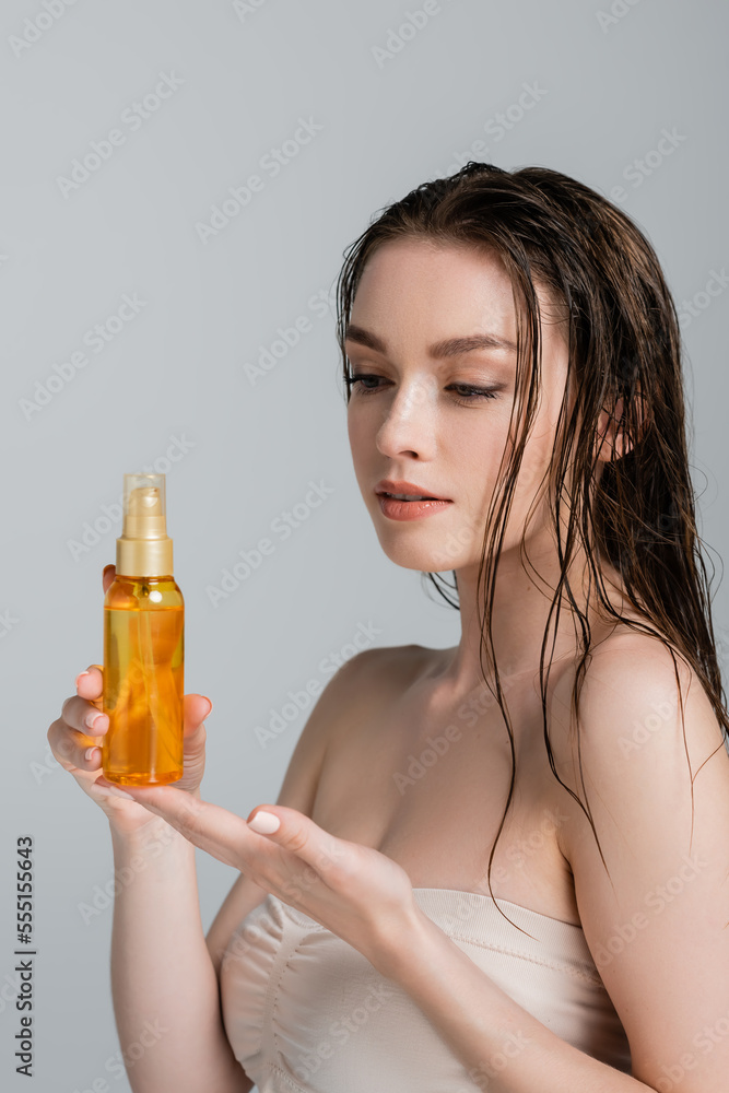 pretty young woman with bare shoulders looking at bottle with hair oil isolated on grey pretty young woman with bare shoulders looking at bottle with hair oil isolated on grey