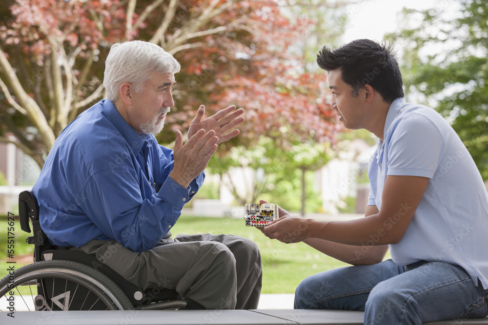 Engineer with muscular dystrophy and diabetes in his wheelchair talking with design engineer about microchips on circuit board Engineer with muscular dystrophy and diabetes in his wheelchair talking with design engineer about microchips on circuit board