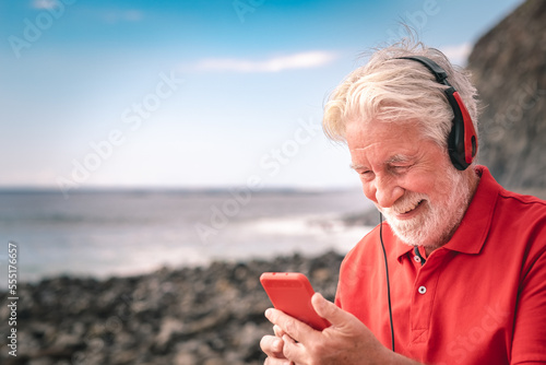 Beautiful senior man in red listening music by headphones standing on the beach - elderly happy man at sea holding smartphone enjoying free time and retirement #555176657