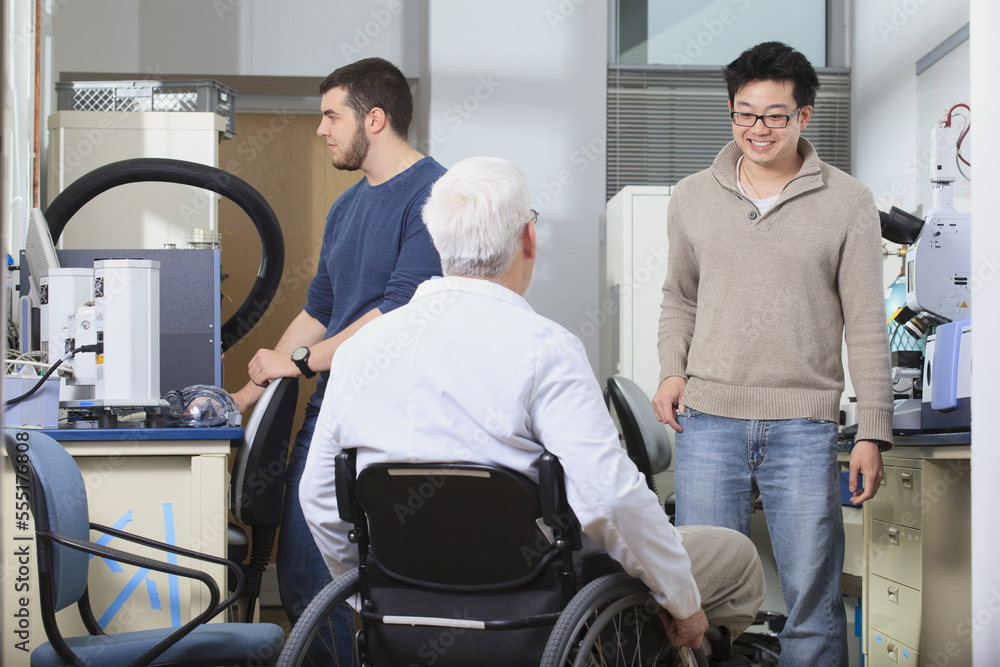Professor with muscular dystrophy talking to engineering students about chemical analysis equipment in a laboratory Professor with muscular dystrophy talking to engineering students about chemical analysis equipment in a laboratory