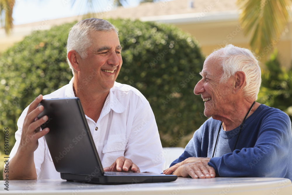 Senior man smiling showing a laptop to his father, who is visually impaired Senior man smiling showing a laptop to his father, who is visually impaired