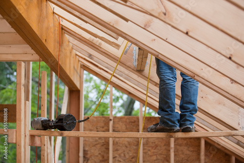 Hispanic carpenter working on roof rafters with nail gun at a house under construction