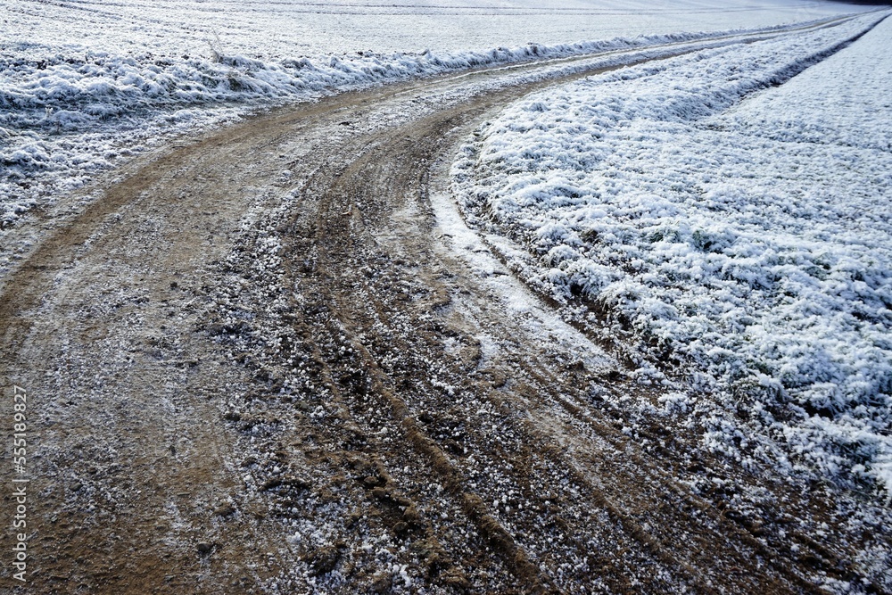 Gefrorener kurviger erdiger Feldweg zwischen Schneebedeckten Feldern bei Frost, Eis und Schnee am Morgen im Winter