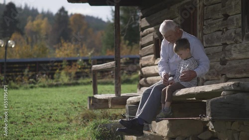 Grandchild and elderly peasant sit together on the porch in front of the rural wooden house