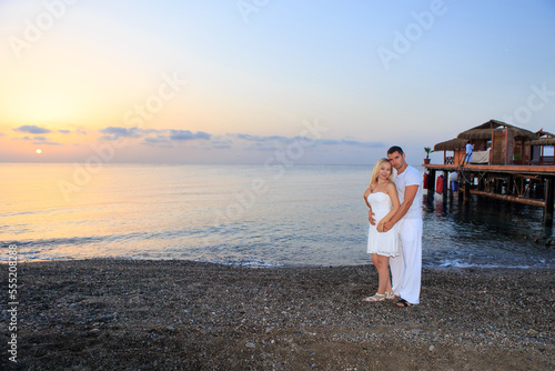 Young couple on the background of the sea sunset or sunrise. Young woman and young brunette man on the sea shore of pebbles.