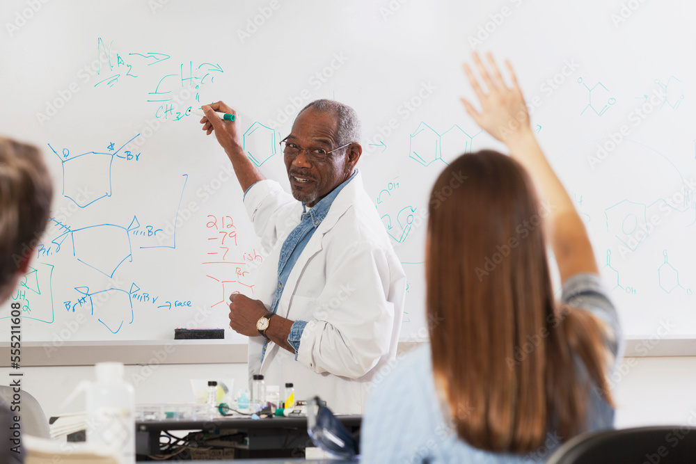 Student raising her hand in an engineering class while professor is at the white board writing about chemical bonds Student raising her hand in an engineering class while professor is at the white board writing about chemical bonds