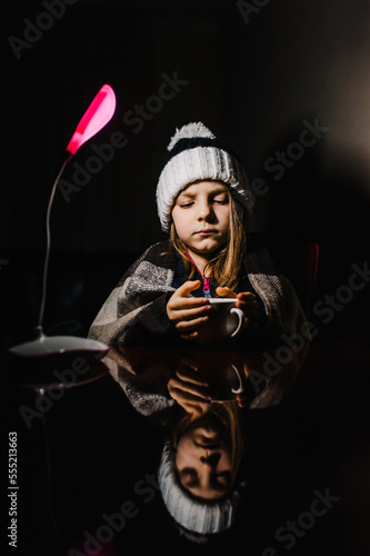 Blackout in Ukraine. A Ukrainian little girl, a child, a schoolgirl in warm clothes, a hat, a blanket, a blanket sits at a table in a cold room in the dark with a lamp and drinks hot tea from a cup.