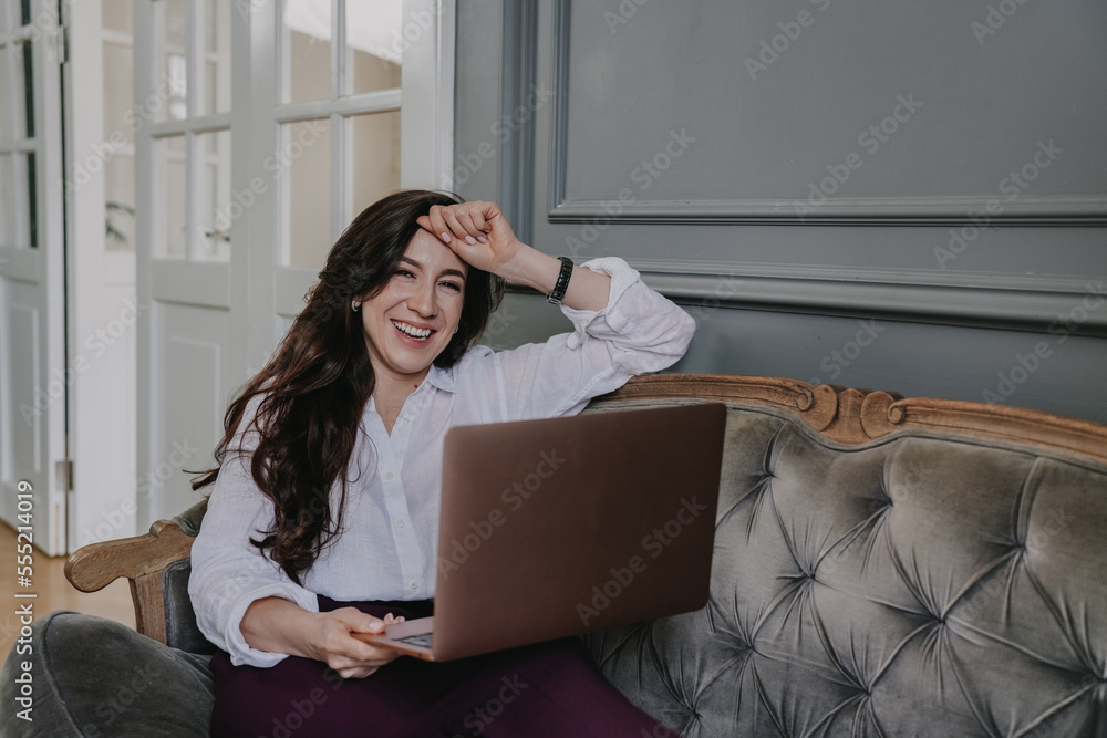 Obraz premium Cheerful Italian brunette woman in white shirt, violet pants sitting on old fashion sofa with laptop looks at camera toothy smiles. Successful hispanic lawyer distant working home. Business, finance.