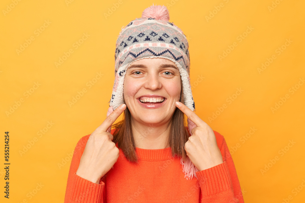 Horizontal shot of delighted young adult woman wearing funny earflap hat and orange casual jumper standing isolated over yellow background, pointing at her teeth after visiting dentist. Horizontal shot of delighted young adult woman wearing funny earflap hat and orange casual jumper standing isolated over yellow background, pointing at her teeth after visiting dentist.