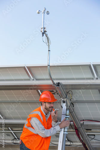 Power engineer on a cell phone while holding weather station conduit at solar photovoltaic array