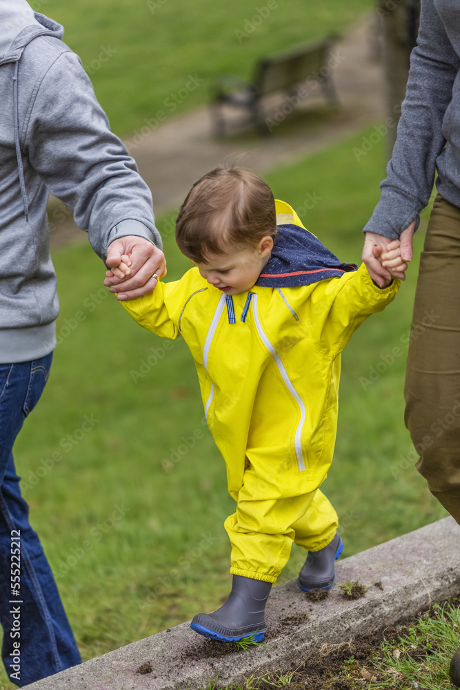 Close-up of mother and father holding hands with young daughter as she walks along concrete ledge in the park; Surrey, British Columbia, Canada Close-up of mother and father holding hands with young daughter as she walks along concrete ledge in the park; Surrey, British Columbia, Canada
