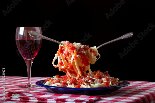 Pasta noodles suspended above a plate of pasta covered with tomato sauce and grated cheese on a red and white checker table cloth with a glass of red wine and napkin; Studio