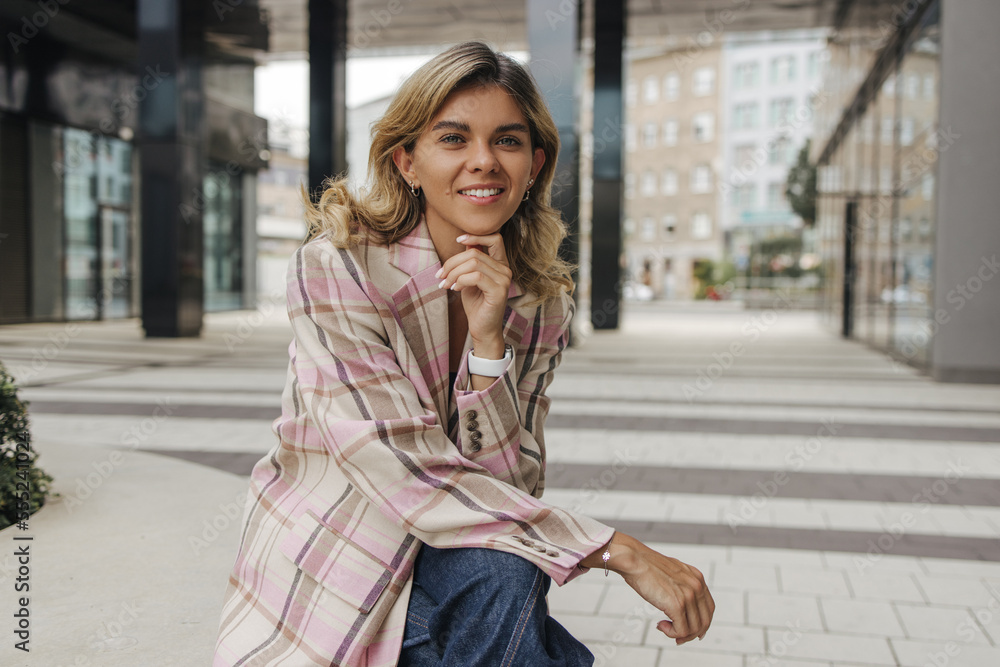 Fototapeta premium Pretty young caucasian woman smiling looking at camera sitting outside office on break. Blonde wears shirt and jeans. Concept of leisure, recreation