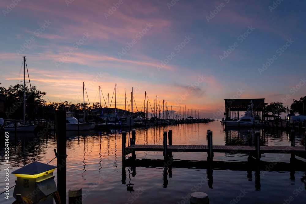 A harbor during the blue hour with silhouettes of the masts of sailboats, a boat house, a pier, and a harbor light with a blue sky and pink clouds.