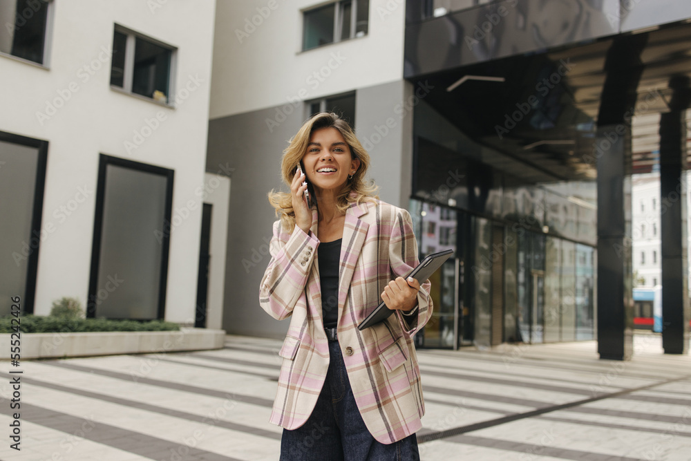 Stylish young caucasian woman looking at camera talking on smartphone outdoors. Blonde looks at camera, wears jacket and jeans. Technology concept, lifestyle